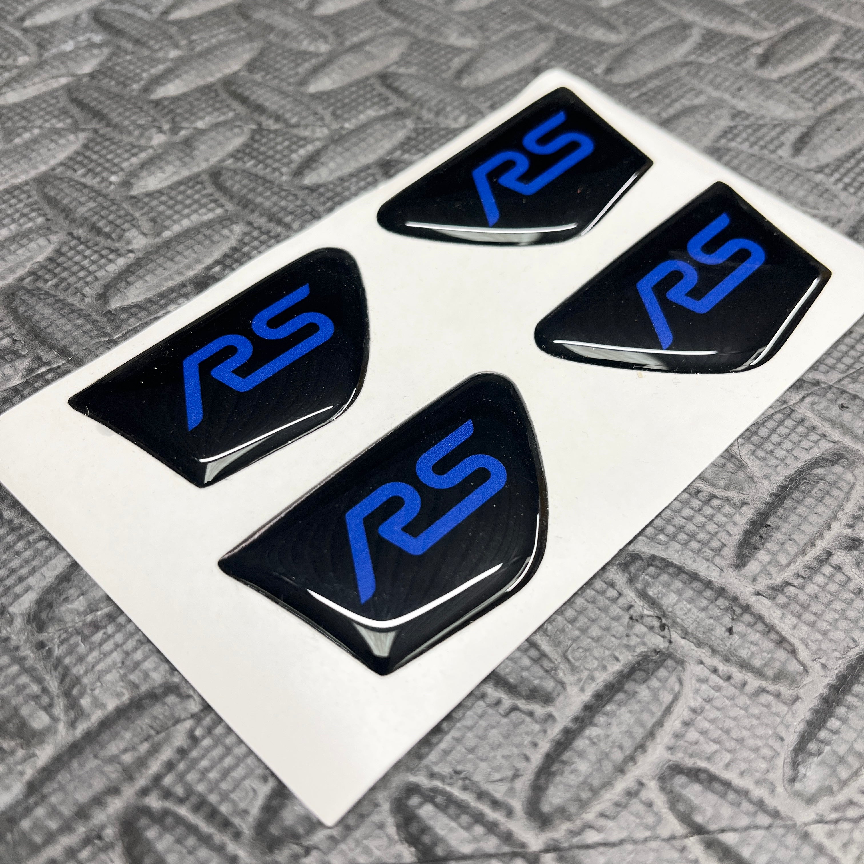 Four black 'RS' logo stickers with blue text on a white backing, placed on a textured gray surface.