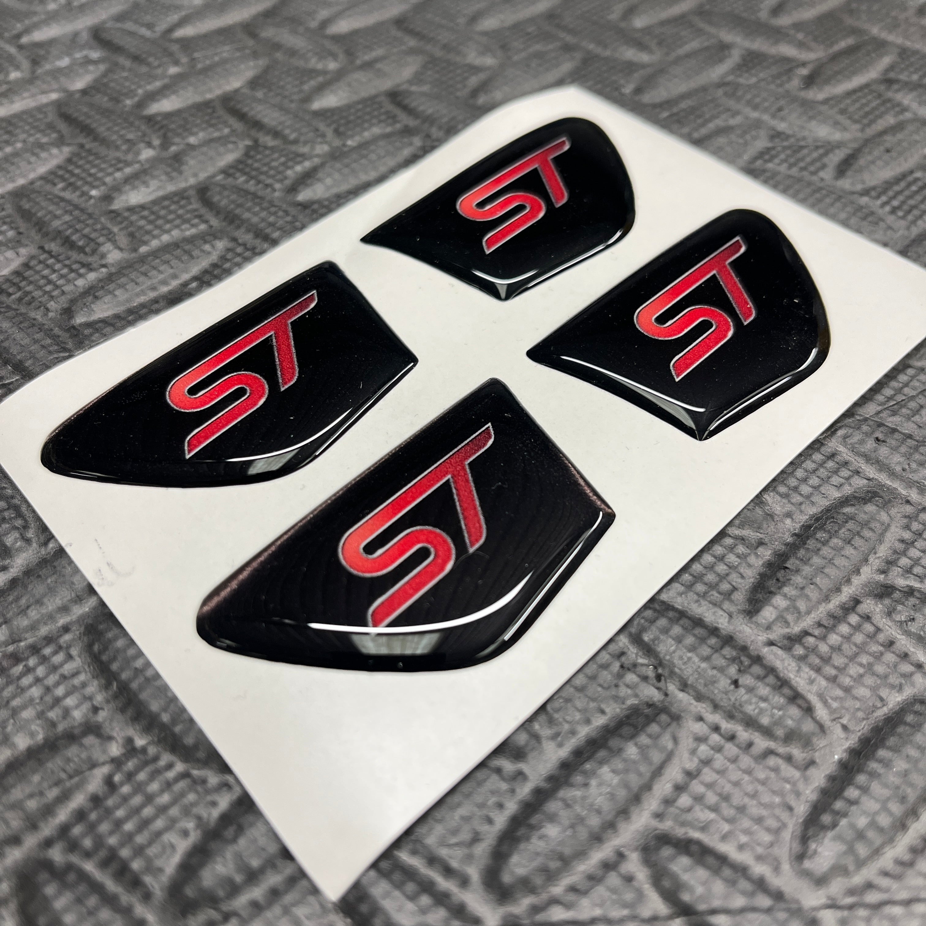 Four black and red 'ST' emblems on a white card with a textured gray background.