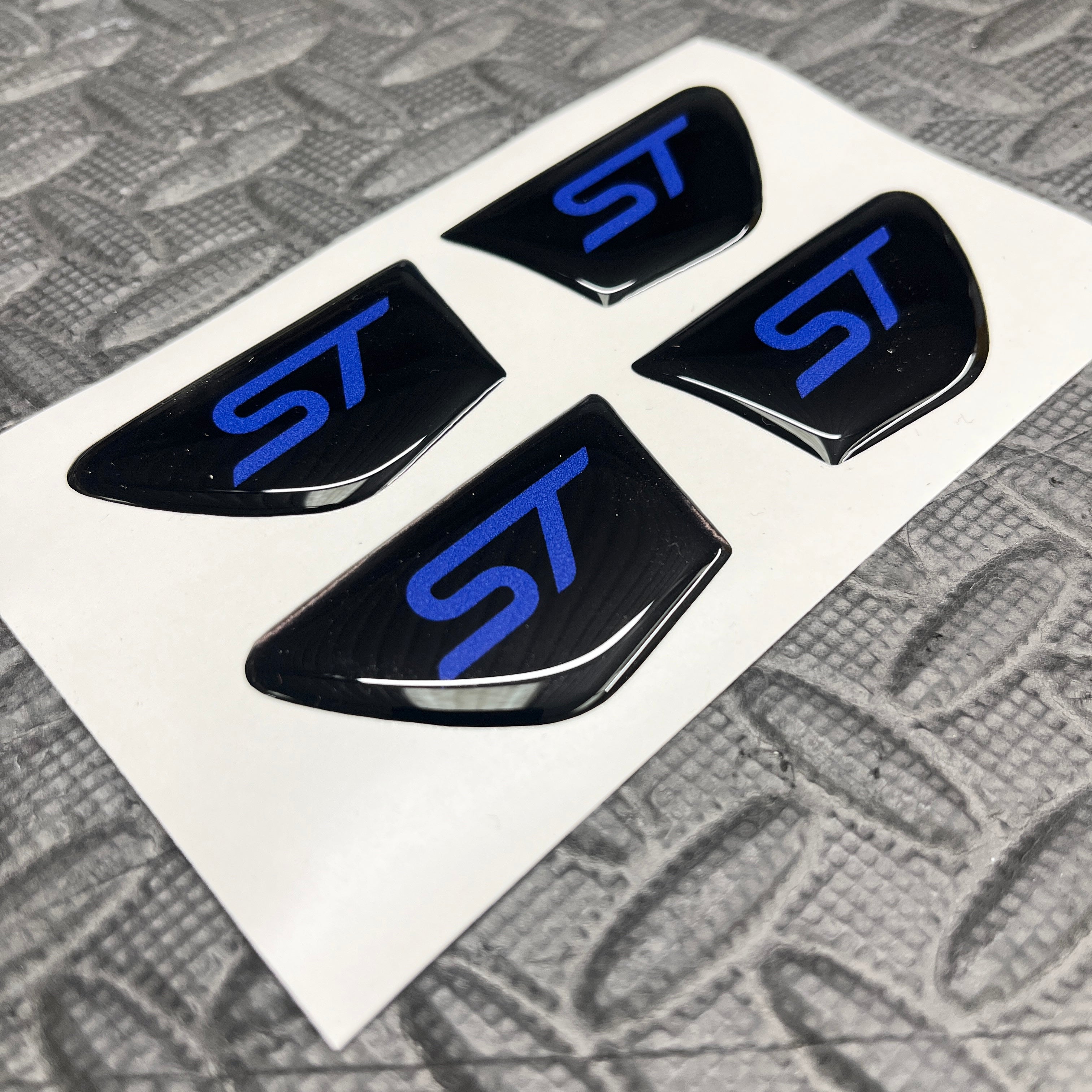 Four black 'ST' logo emblems on a white card with a textured gray background