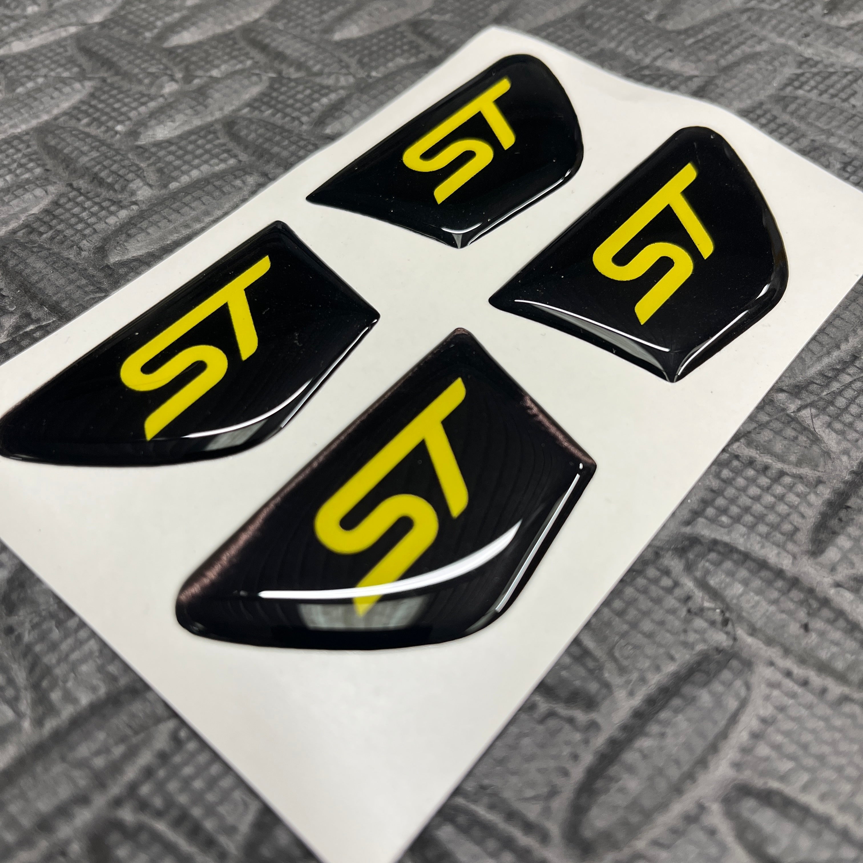 Four black and yellow 'ST' logo stickers on a white card with a textured gray background.