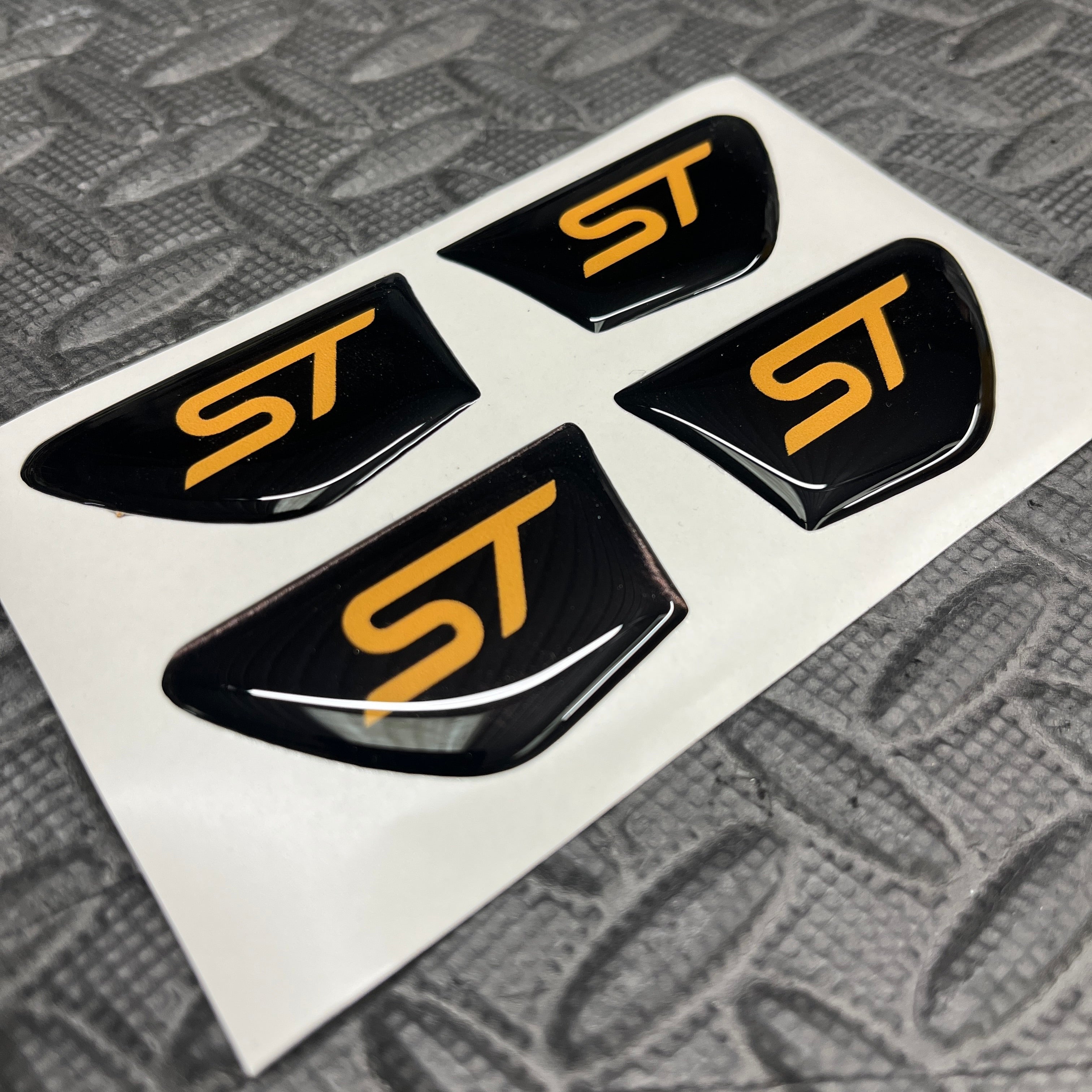 Four black and gold 'ST' logos on a white card with a textured gray background