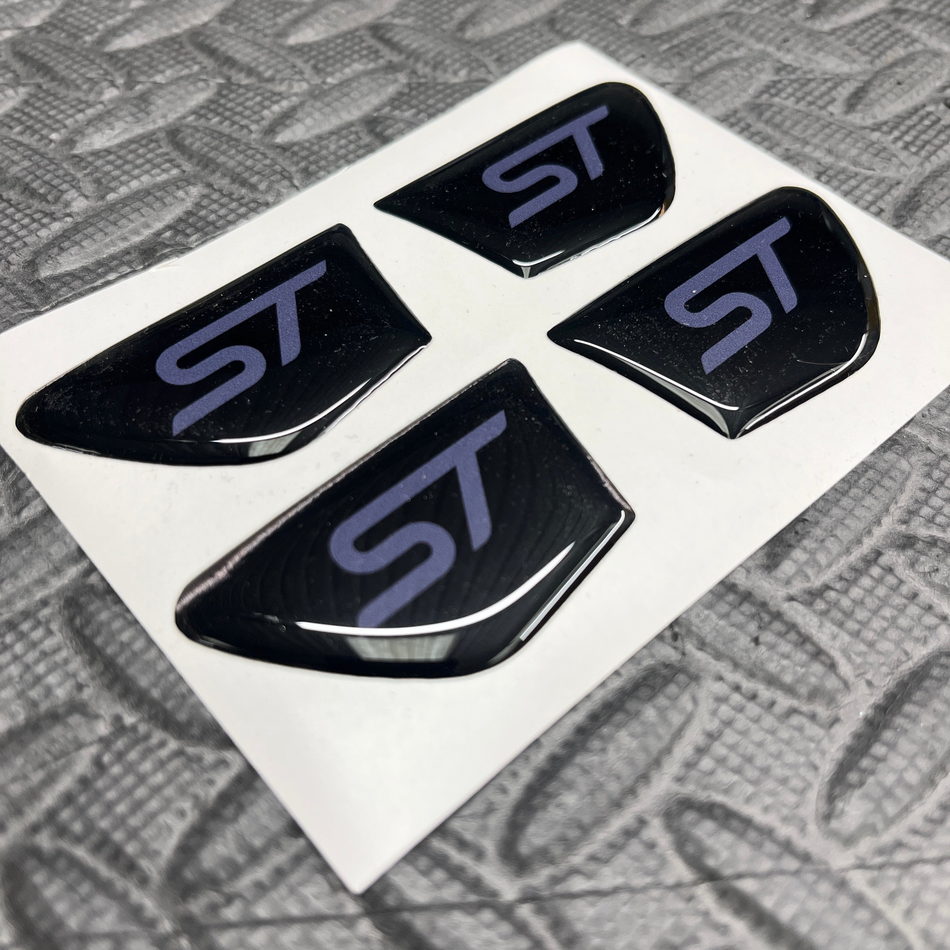 Four black 'ST' logo car emblems on a white card with a textured gray background.