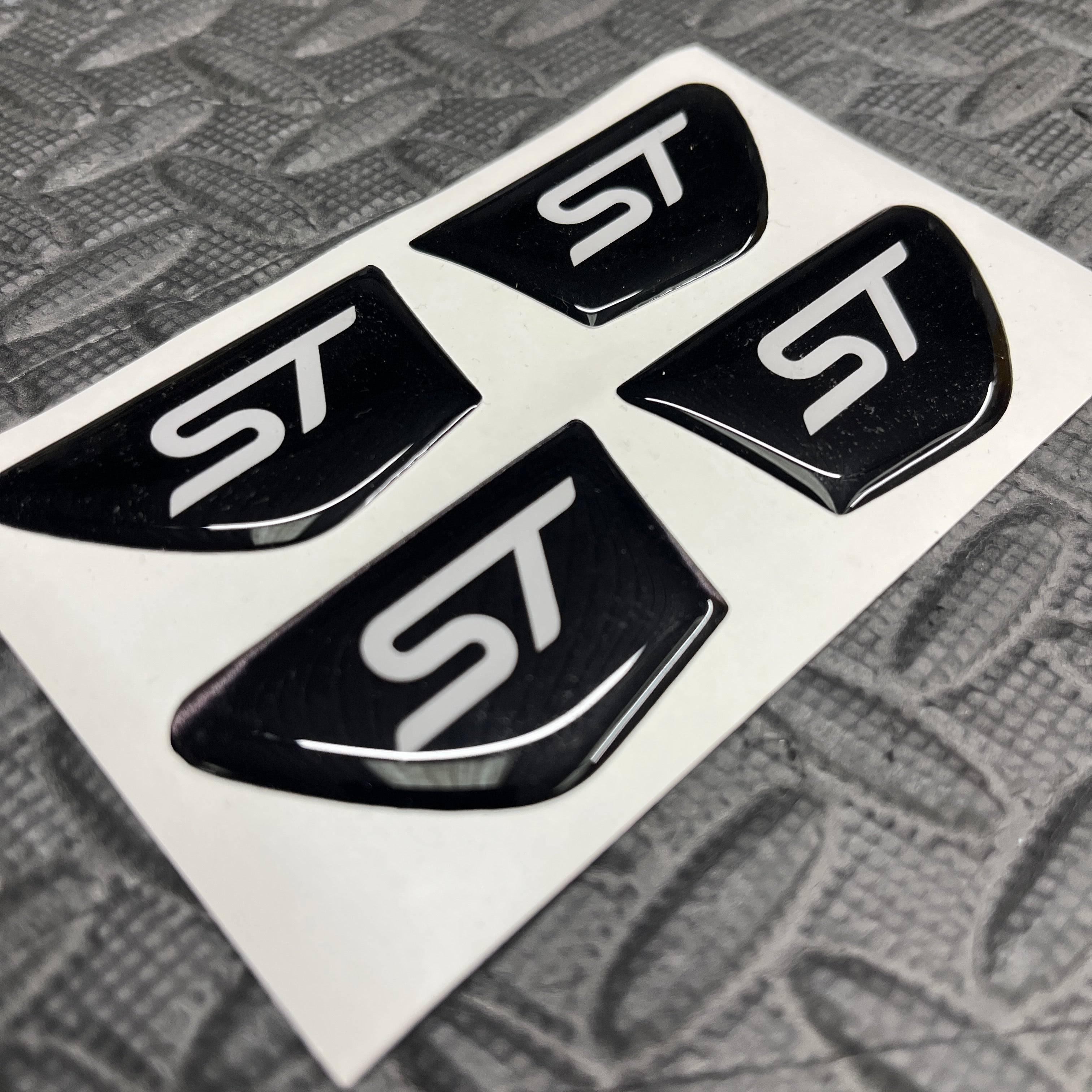 Four black 'ST' logos on a white backing with a textured gray surface below.